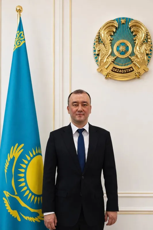 Жанболат Усенов стоит возле флага и герба Казахстана